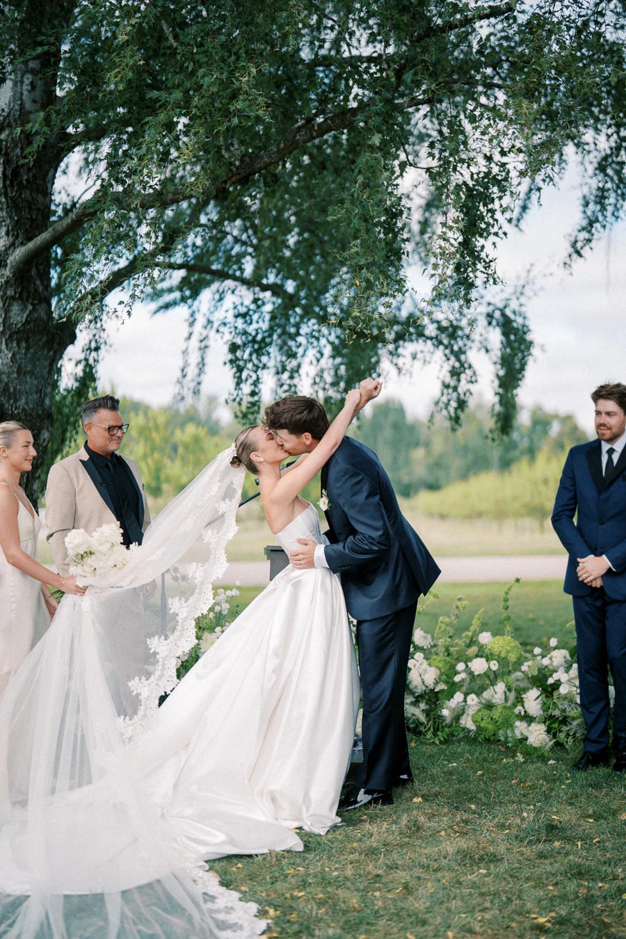 Bride and groom sharing a kiss during outdoor wedding ceremony under a tree, surrounded by bridal party, with lush greenery and floral decorations.