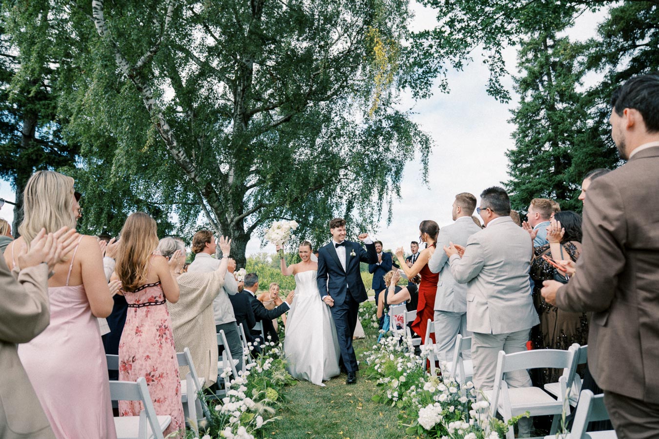 Newlywed couple walking down the outdoor wedding aisle, surrounded by cheering guests and lush greenery.