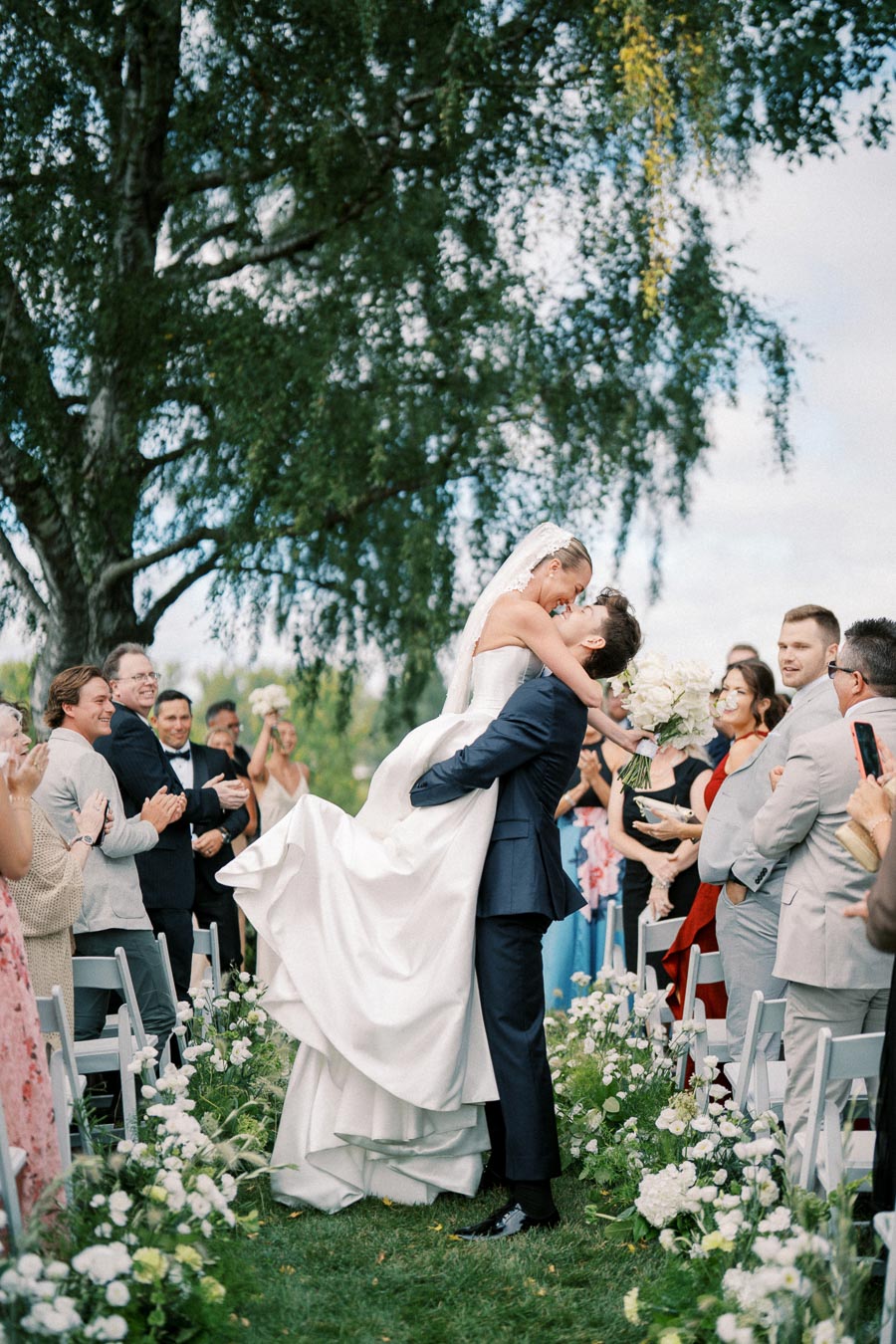 A bride and groom embrace joyfully at an outdoor wedding ceremony, surrounded by applauding guests seated beside white floral arrangements, with lush green trees in the background.