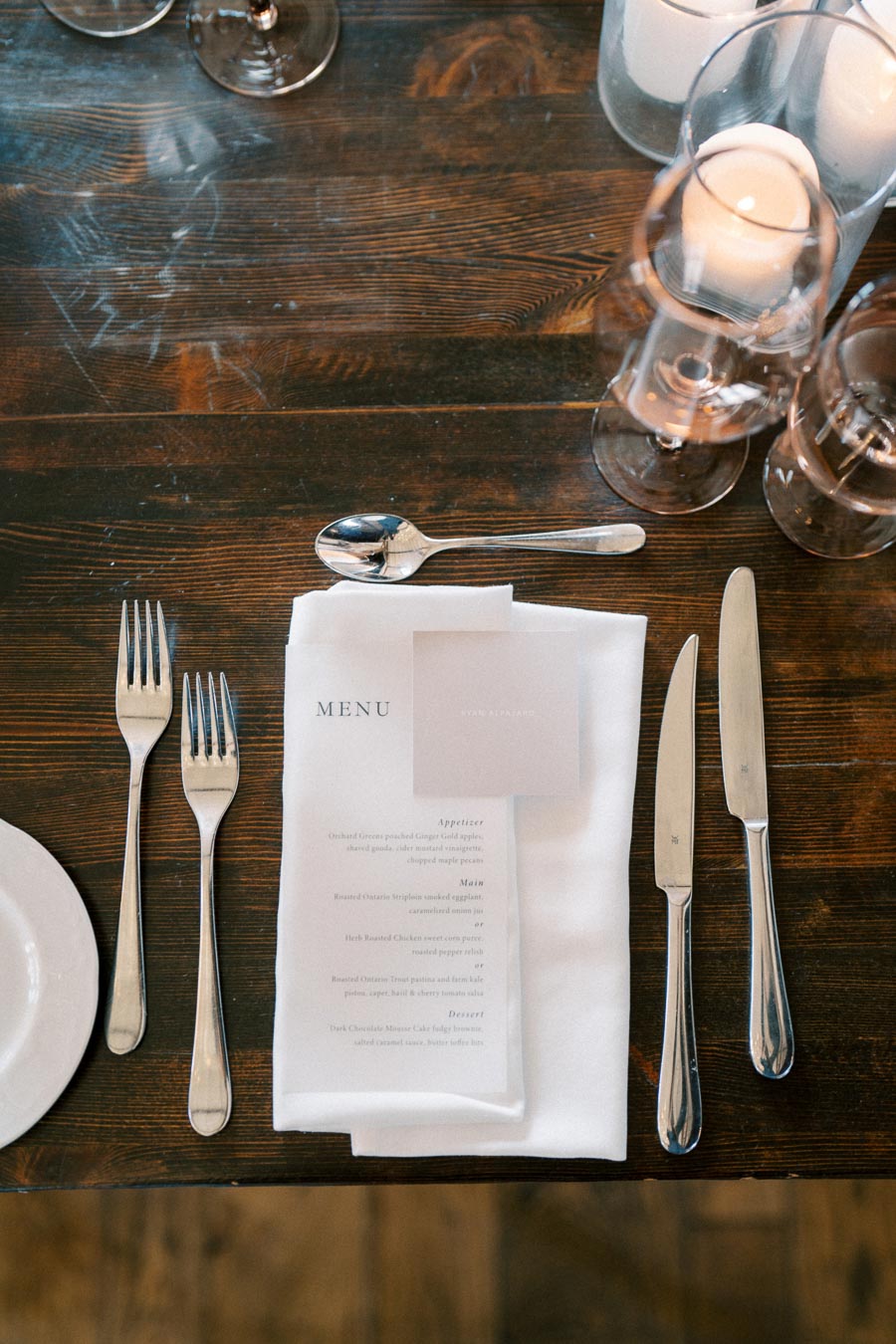 Elegant dining setup with a wooden table, featuring a white napkin adorned with a printed menu card. Cutlery includes two forks, a spoon, and a knife neatly arranged at each side, accompanied by clear glassware, creating a sophisticated ambiance.