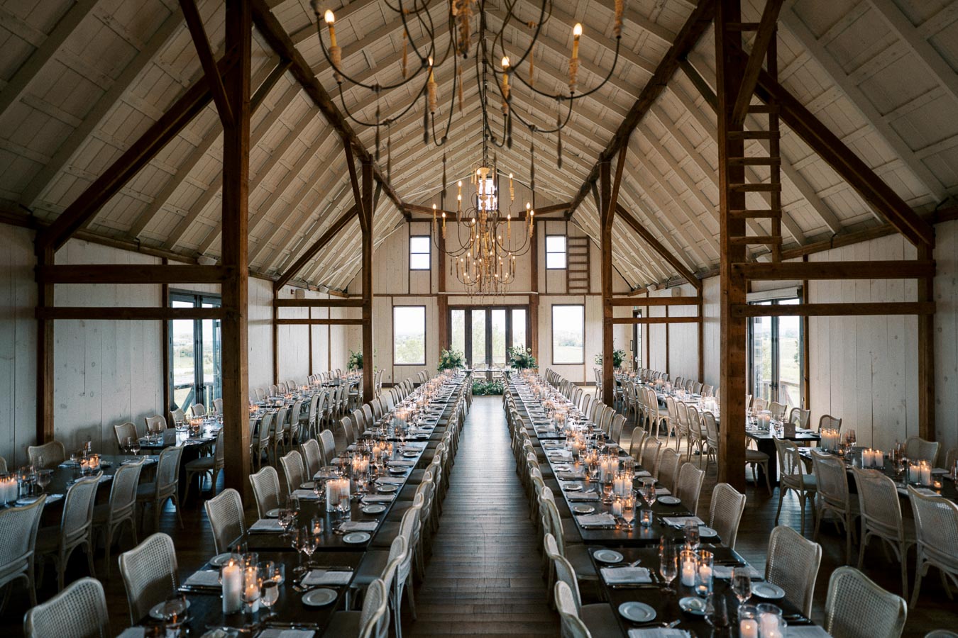 Spacious rustic barn venue with wooden beams and high ceilings, elegantly decorated for a formal event. Long dining tables are set with candles, glassware, and white plates, ready for guests. Natural light streams through large windows, highlighting the charming ambiance.