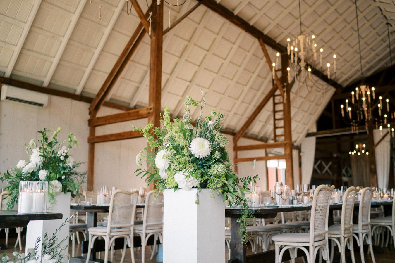 Elegant barn wedding venue with rustic wooden beams, adorned with lush green and white floral centerpieces on white pedestals. The dining area features beautiful candlelit tables set with fine tableware, surrounded by vintage wooden chairs, under chandeliers for a romantic atmosphere.