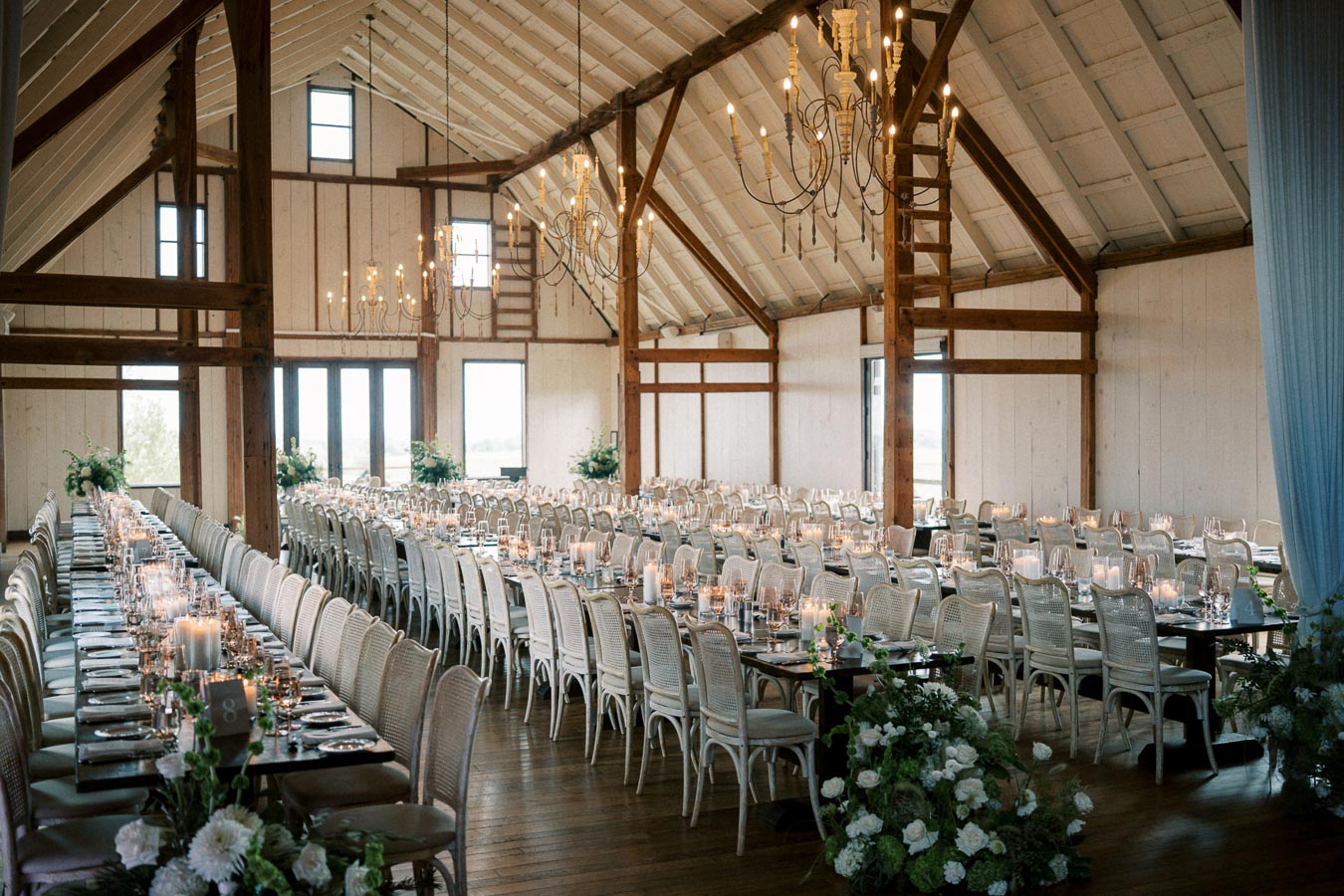 Elegant barn wedding reception setup with long dining tables, glowing candles, and rustic chandeliers in a spacious venue.