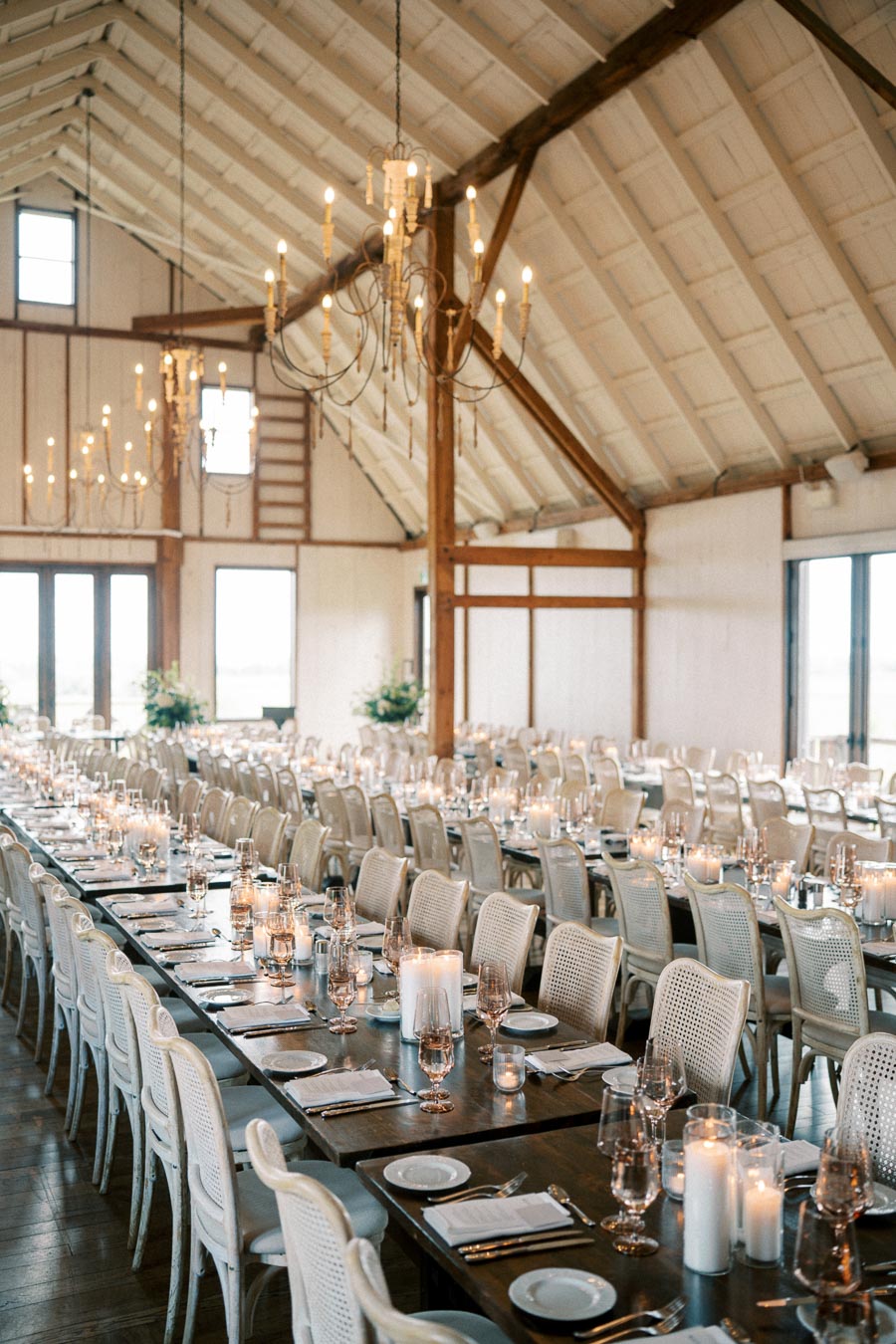 Elegant wedding reception setup in a rustic barn venue, featuring long banquet tables with white chairs, adorned with candles and glassware. Chandelier lighting, wooden beams, and high ceilings add a charming ambiance, ready for a festive celebration.