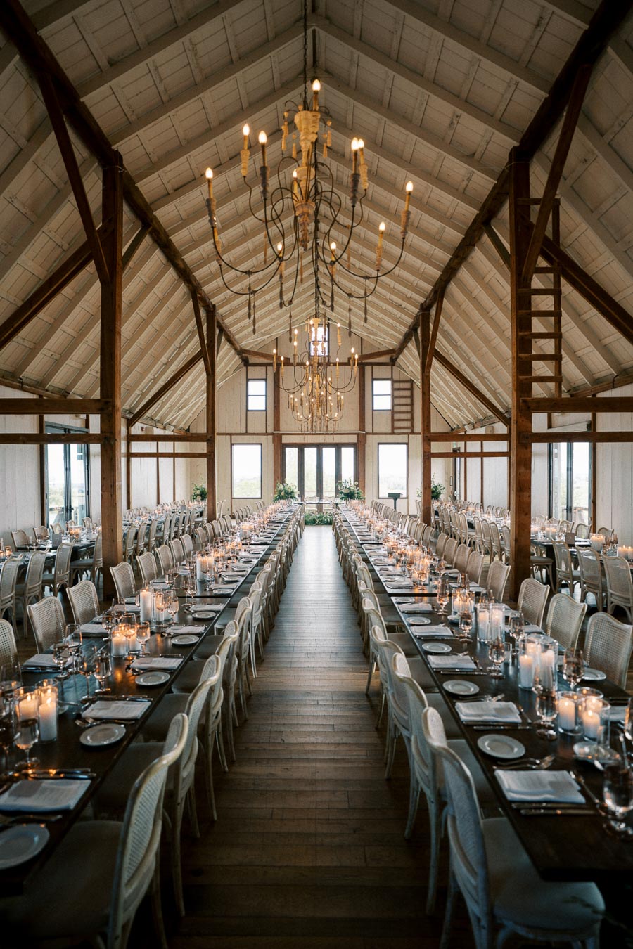 Elegant barn wedding reception setup with long wooden tables decorated with candles and place settings under rustic chandeliers.