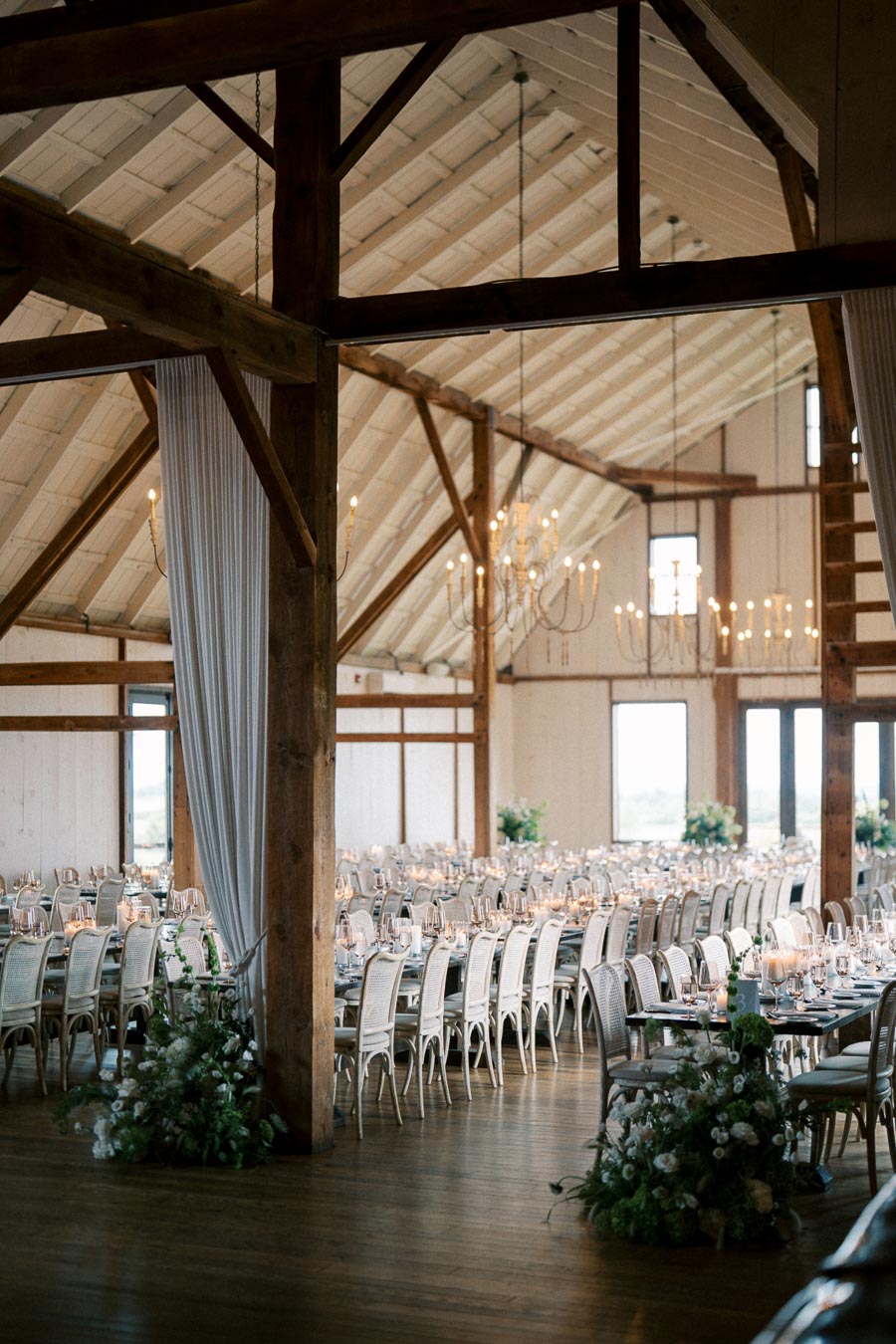 Alt text: Elegant rustic wedding reception setup in a barn venue with long banquet tables, white chairs, floral centerpieces, and ambient lighting from chandeliers.
