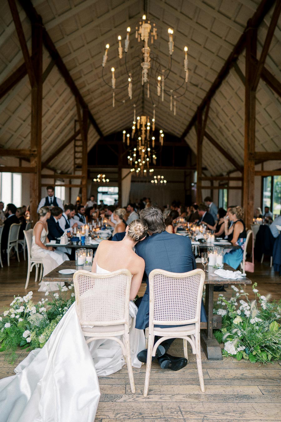 Elegant wedding reception in rustic barn with bride and groom seated closely, adorned with chandeliers and floral arrangements.