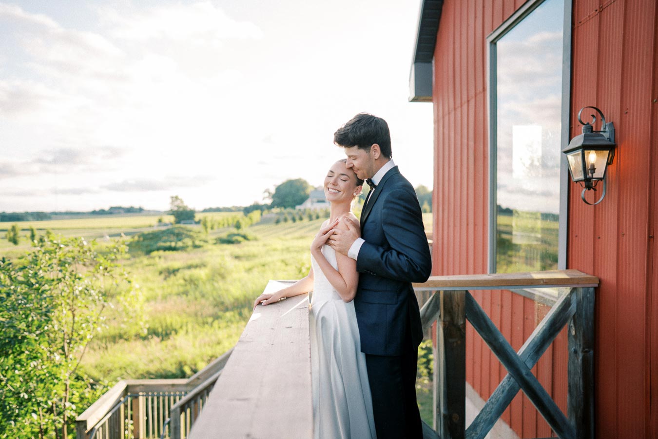 Romantic couple embracing on a sunny balcony overlooking lush green countryside, next to a red building.