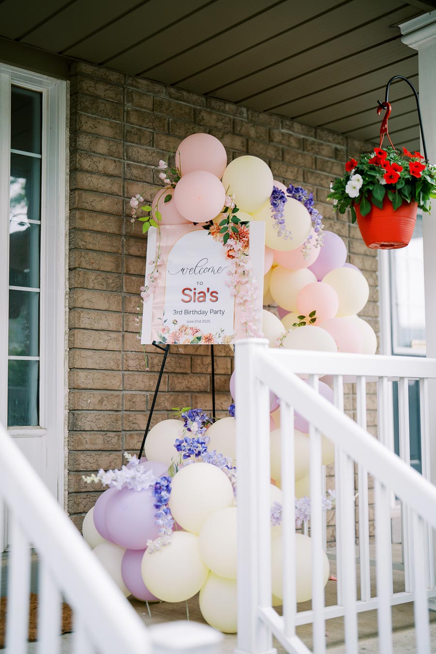 Colorful balloon arch with a welcome sign for a 3rd birthday party on a porch, surrounded by pink, purple, and yellow decorations, with a potted plant nearby.