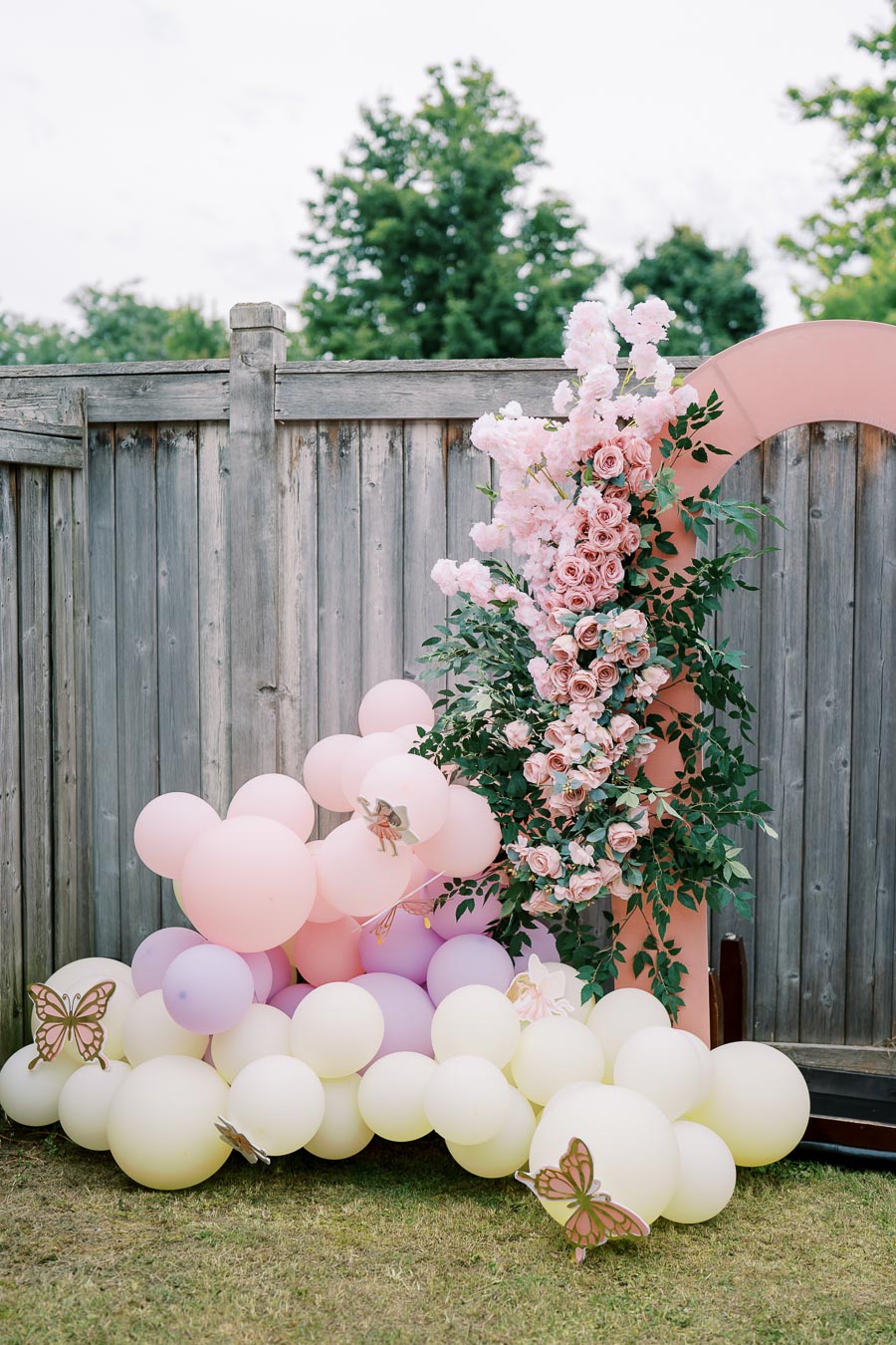 Pastel balloon arrangement with pink and white flowers against a wooden fence, featuring decorative butterflies, ideal for garden celebrations and parties.