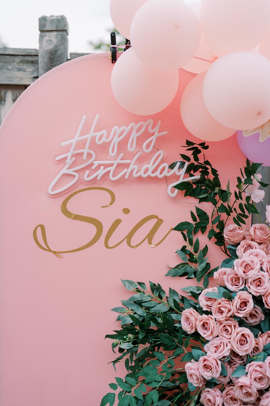 Pink birthday backdrop with Happy Birthday Sia text, adorned with pink balloons and a bouquet of lush pink roses and green leaves.