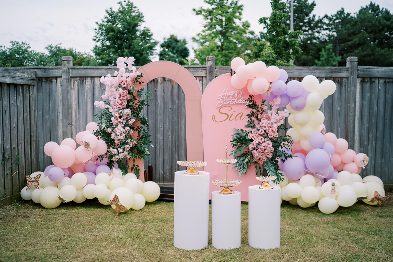 A beautifully decorated outdoor birthday setup featuring a pink and pastel balloon garland, floral arrangements with pink roses, and stylish gold tiered stands on white pedestals. The backdrop includes a personalized birthday message. Perfect for celebrations and event inspiration.