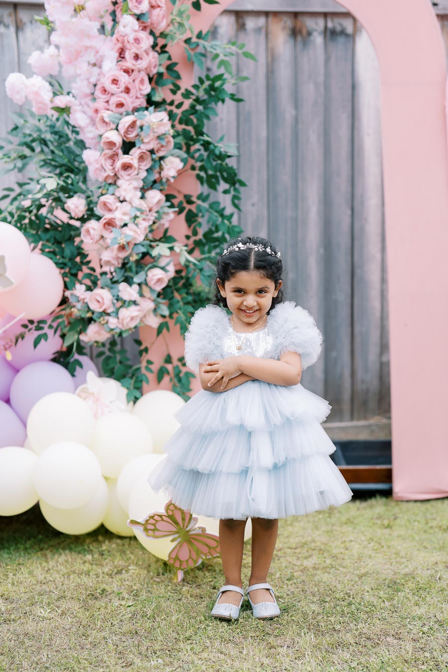 Young girl in a light blue ruffled dress standing outdoors, surrounded by pastel pink and purple balloons and floral decorations, smiling at a party or celebration event.