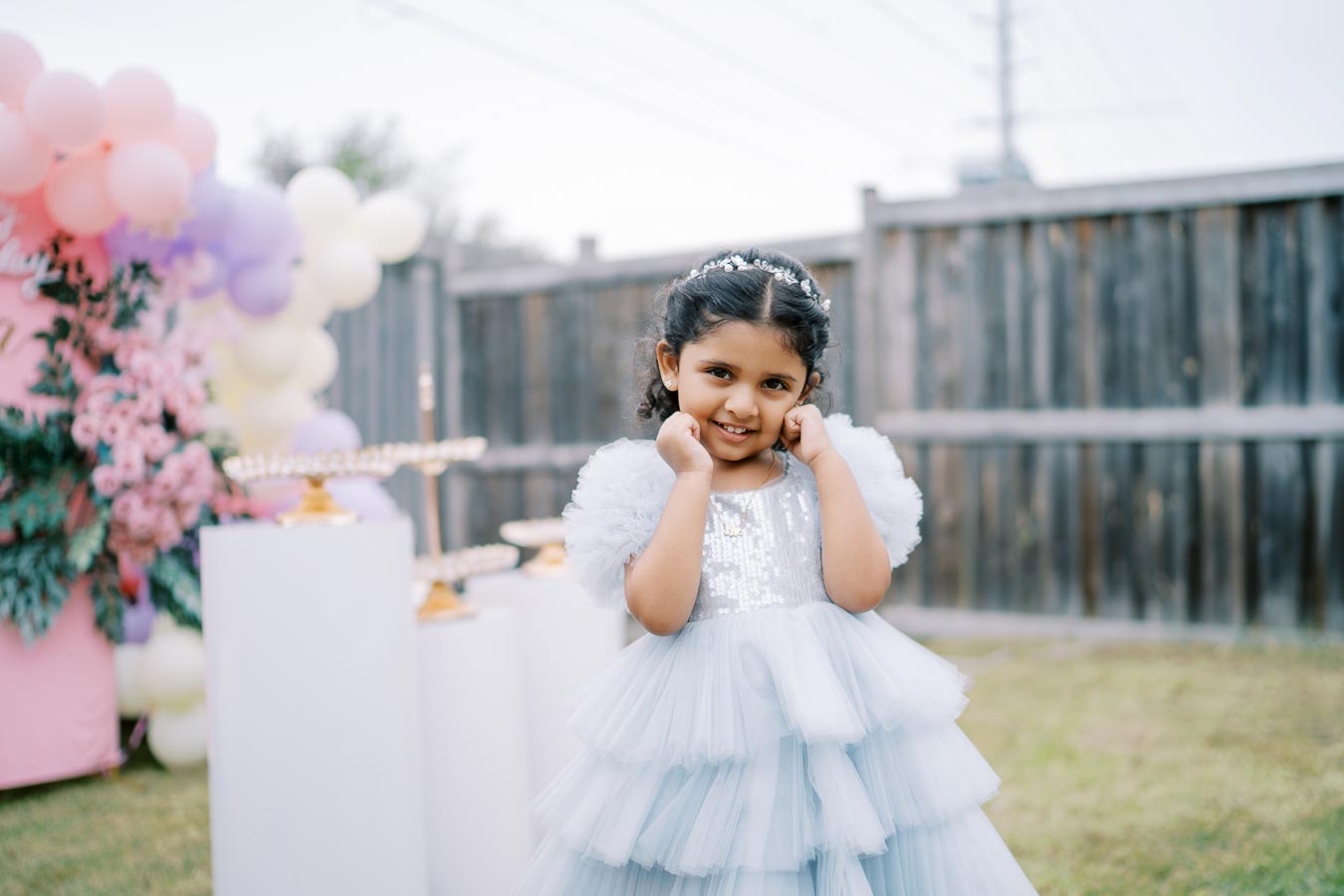 A young girl wearing a fluffy blue dress poses happily at an outdoor party with a backdrop of colorful balloons and decorations.