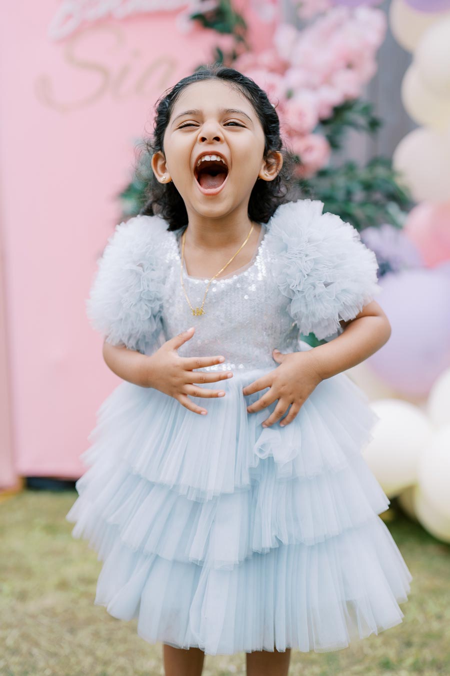 A joyful young girl in a fluffy light blue dress, laughing with excitement at an outdoor celebration decorated with pink and white balloons.