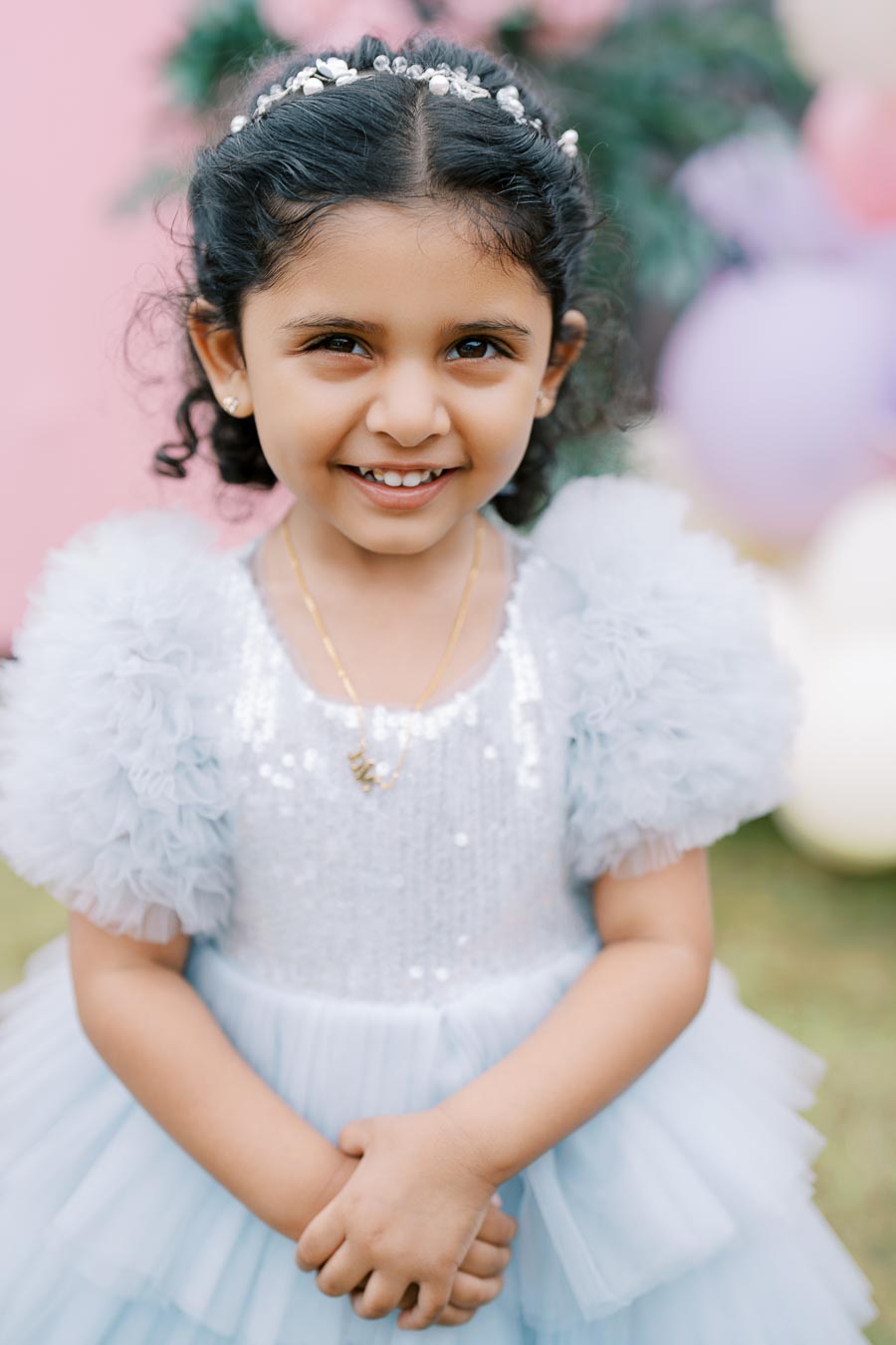 Smiling young girl in a light blue, ruffled dress with a sequin top, standing outdoors against a pink and green blurred background, wearing a beaded headband and a necklace.