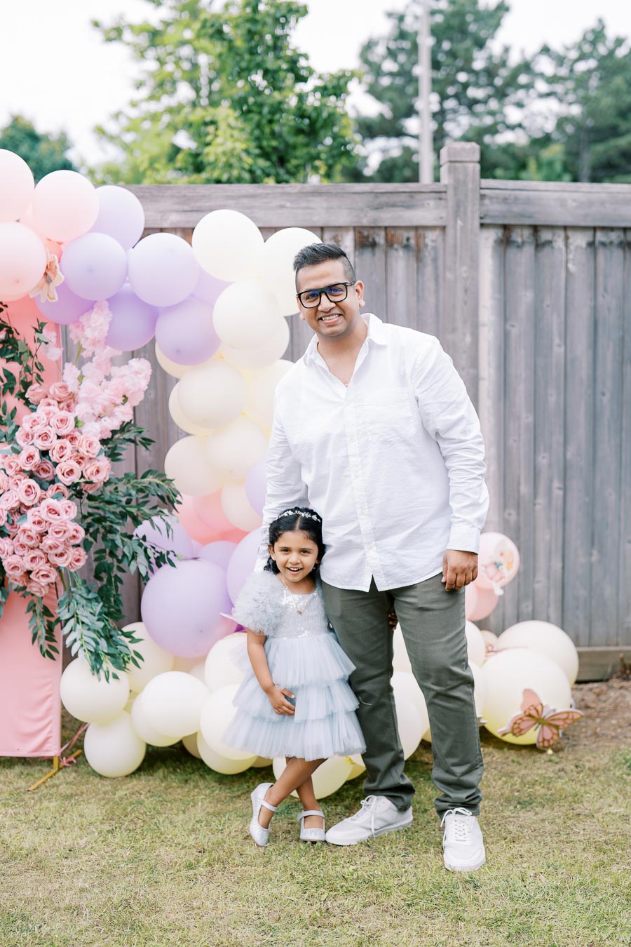 Father and daughter smiling at a garden party, with a colorful balloon arch and floral decorations in the background.