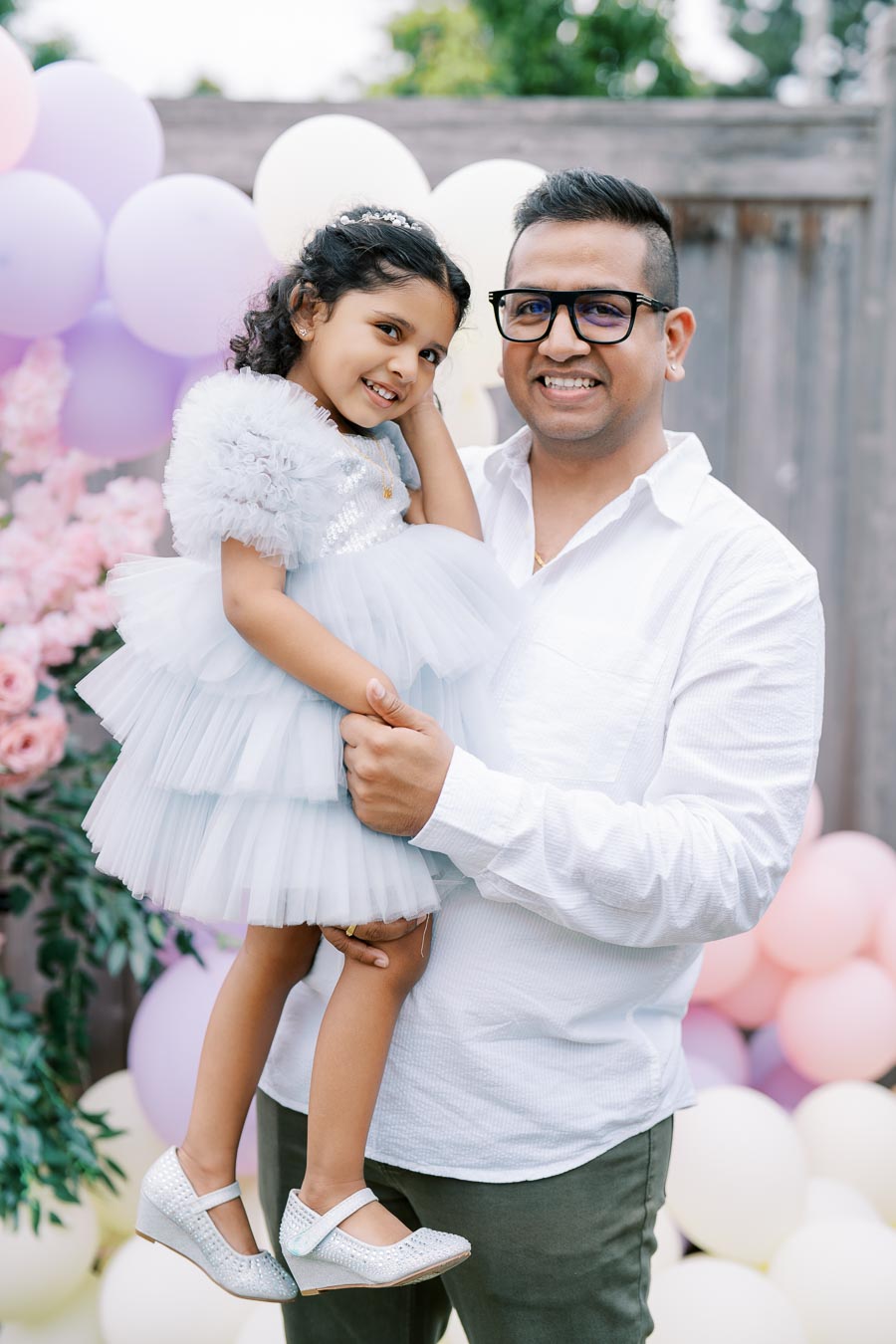 Father in a white shirt holding his smiling daughter in a fluffy blue dress, surrounded by pastel balloons and pink flowers at a celebration.