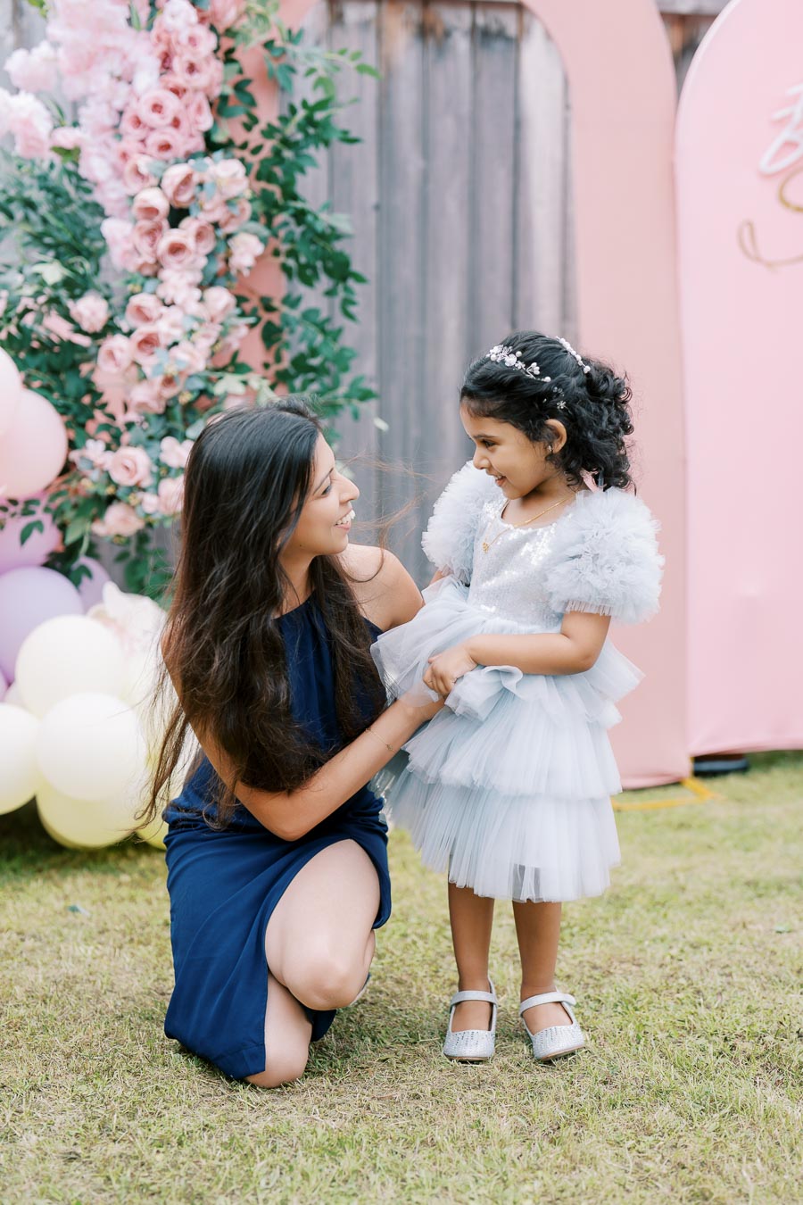 A mother kneeling and smiling at her daughter in a fluffy blue dress, surrounded by pink flowers and balloons in a garden setting.