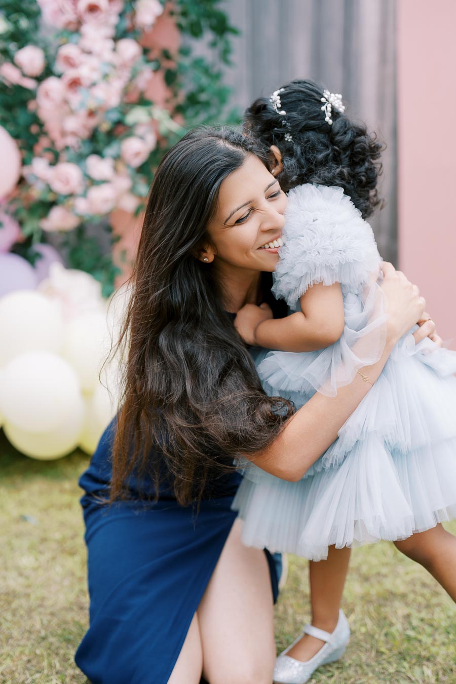A woman joyfully hugging a child in a fluffy dress at an outdoor celebration, with a floral background and pastel balloons.