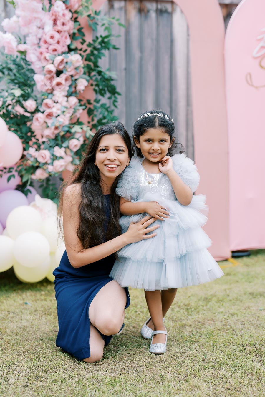 A woman in a blue dress kneeling beside a young girl in a fluffy light blue dress decorated with sequins and a matching hairband, posing happily in front of a pink floral backdrop with balloons at an outdoor event.