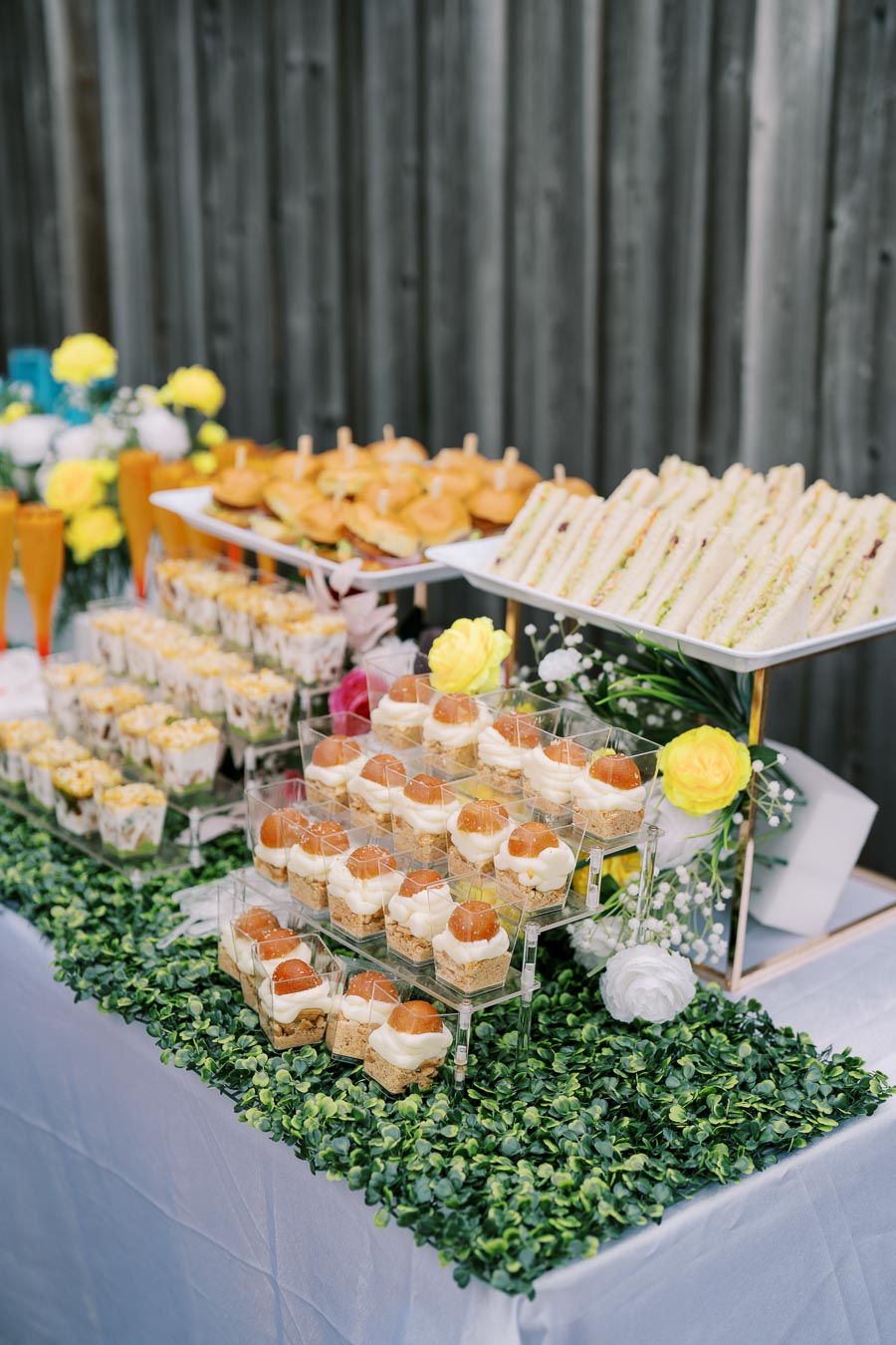 A beautifully arranged buffet table featuring an assortment of finger foods, including mini sandwiches, sliders, and desserts in glass cups, accented by decorative flowers and greenery.