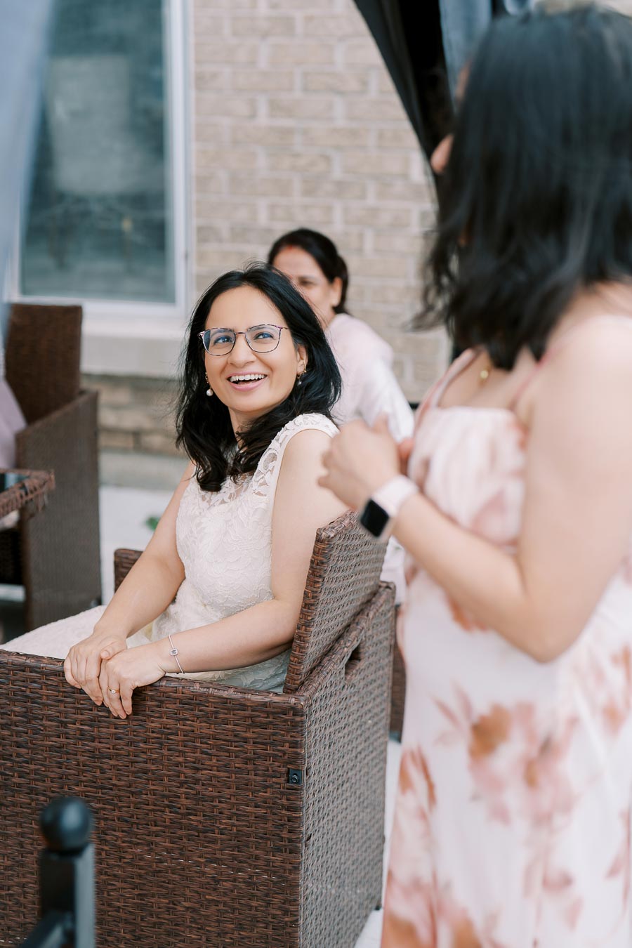 Smiling woman seated in a wicker chair outdoors, interacting with another person, with a brick wall and window in the background.