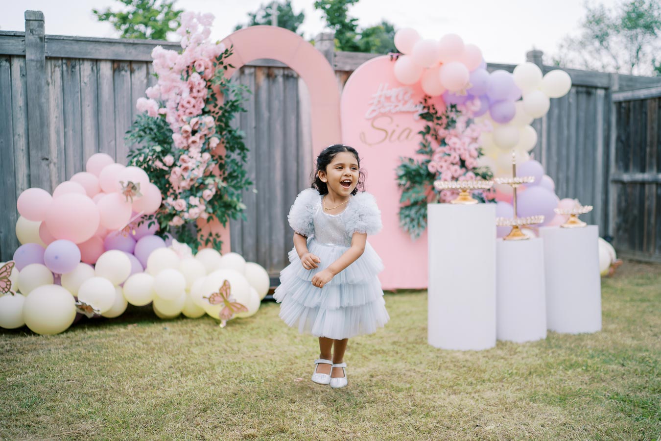 A young girl in a light blue, tiered dress joyfully celebrates at a garden birthday party. She stands in front of a pink and purple balloon and floral backdrop, with decorative pillars. The festive setting includes pastel-colored balloons and floral arrangements, creating a cheerful atmosphere.