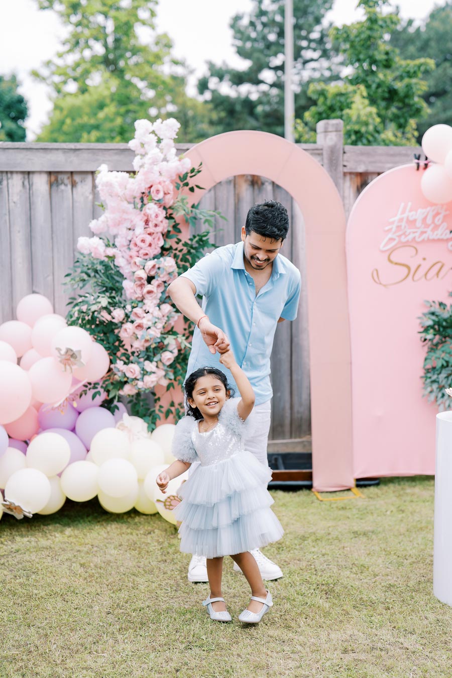 Father and daughter dancing joyfully at an outdoor birthday party, surrounded by pink and white balloons and floral decorations, with a festive Happy Birthday sign in the background.