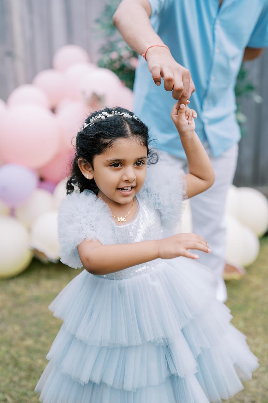 A young girl in a light blue ruffled dress dances joyfully with a smiling adult in a pastel-themed outdoor celebration, surrounded by colorful balloons and greenery.