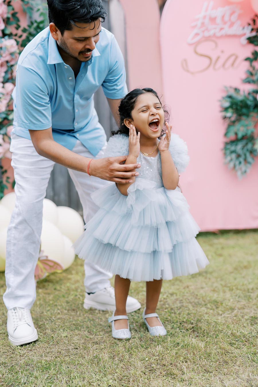 Father and young daughter at an outdoor birthday party, with the child in a blue dress laughing joyfully in front of a pink birthday sign.