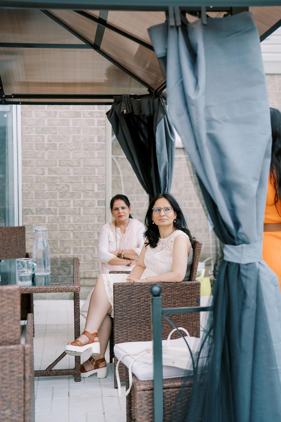 Two women seated in an outdoor patio area, surrounded by wicker furniture and soft curtains. One woman is wearing a white dress and glasses, while the other is dressed in light attire. A glass pitcher and mug are placed on the glass table nearby. Brick wall and canopy structure provide a cozy outdoor setting.