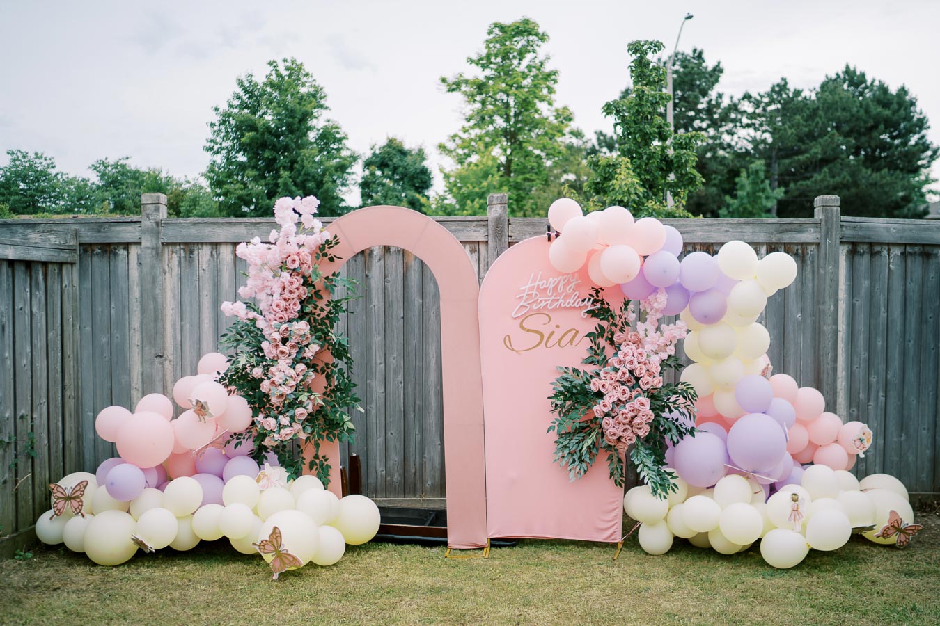 Outdoor birthday party backdrop featuring pastel-colored balloon arches and lush pink floral arrangements set against a wooden fence, with Happy Birthday Sia written in elegant script.