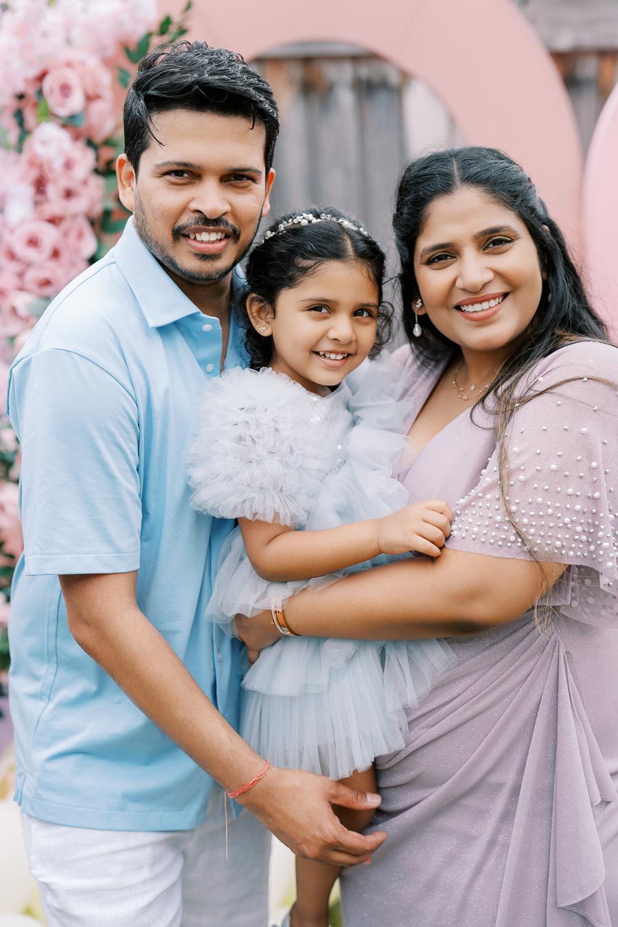 A happy family posing together at an outdoor celebration with a backdrop of pink flowers and balloons, featuring a father in a light blue shirt, a mother in a lavender dress, and a young daughter in a fluffy light blue dress.