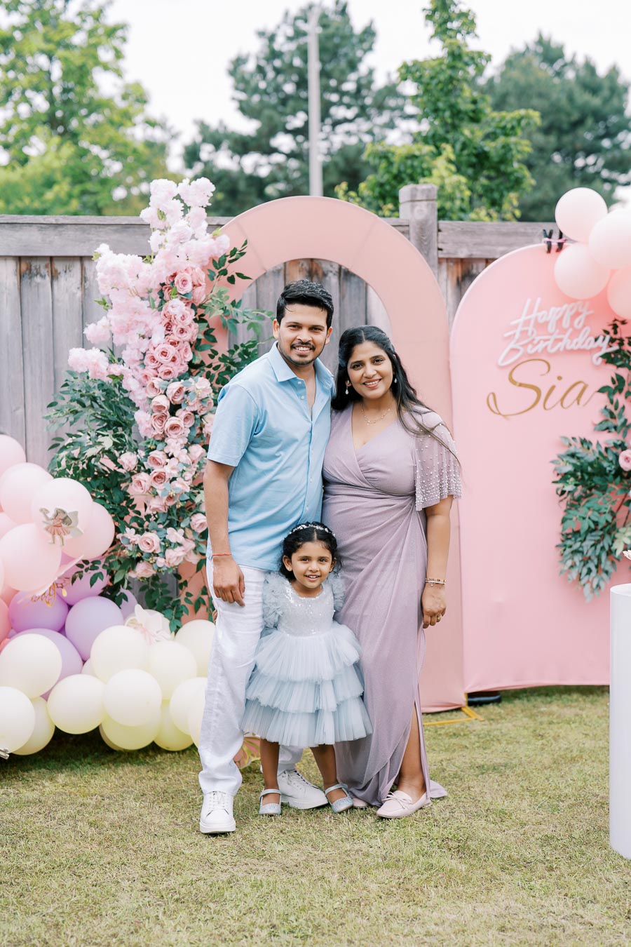 Family celebrating a young girl's birthday in a garden setting adorned with pink floral decorations and balloons.