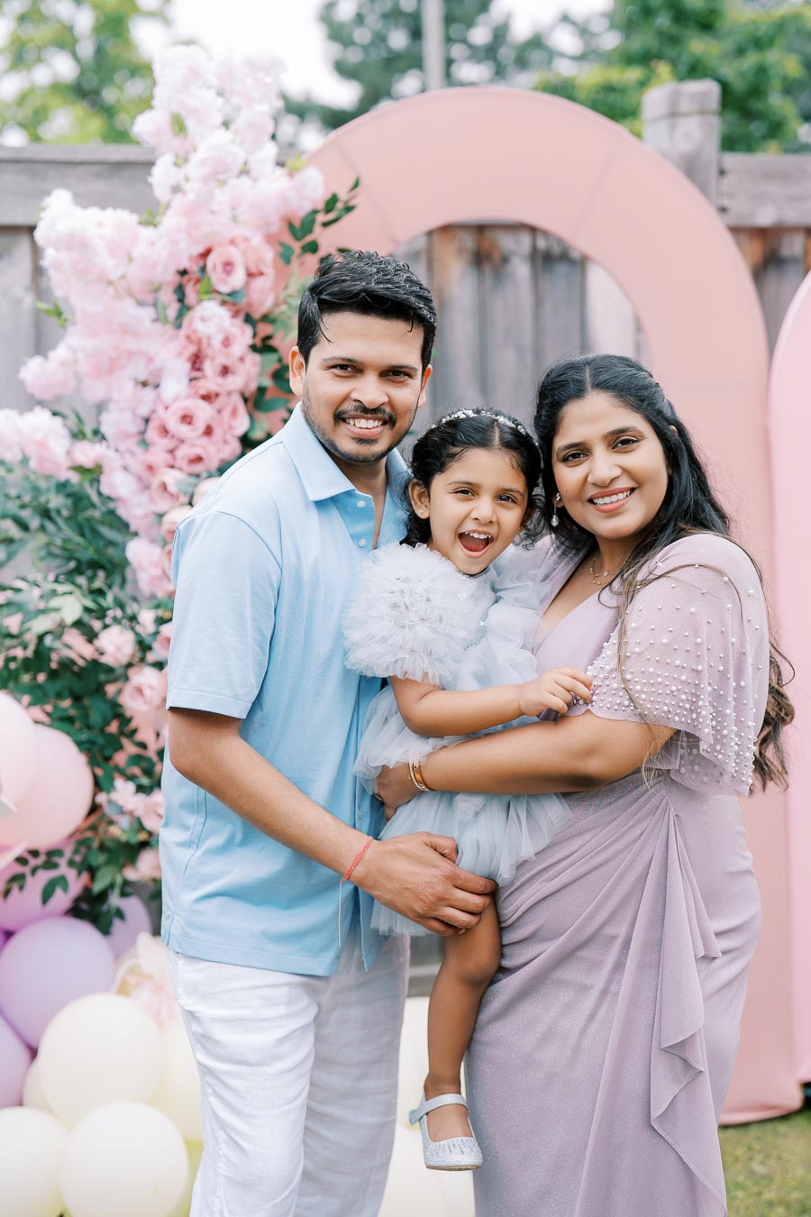 Family celebrating outdoors with balloons and floral decorations, featuring a smiling couple with their young daughter in a light blue dress.