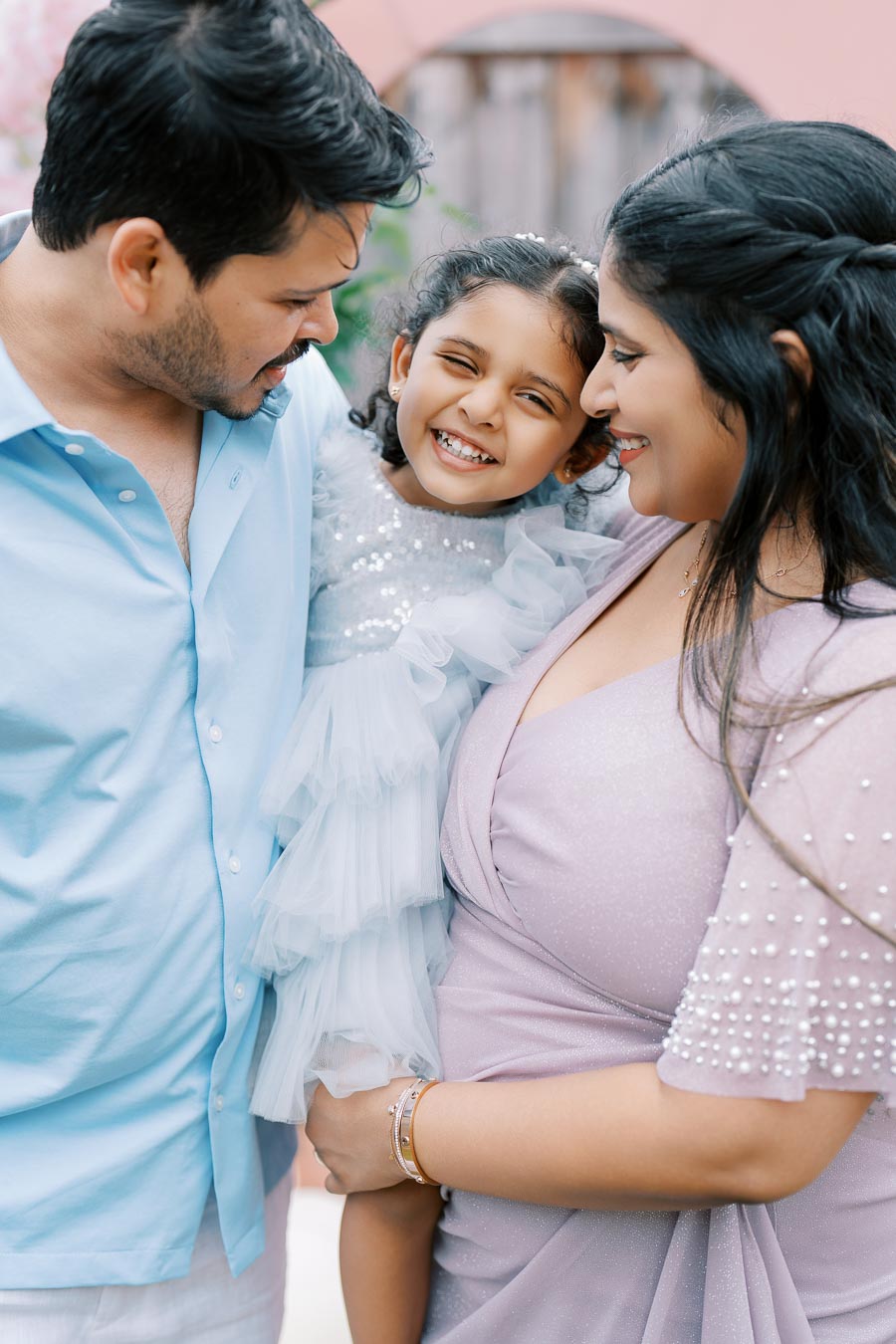 Smiling family embracing outdoors, with a young girl in a ruffled dress between her parents, capturing a joyful moment.
