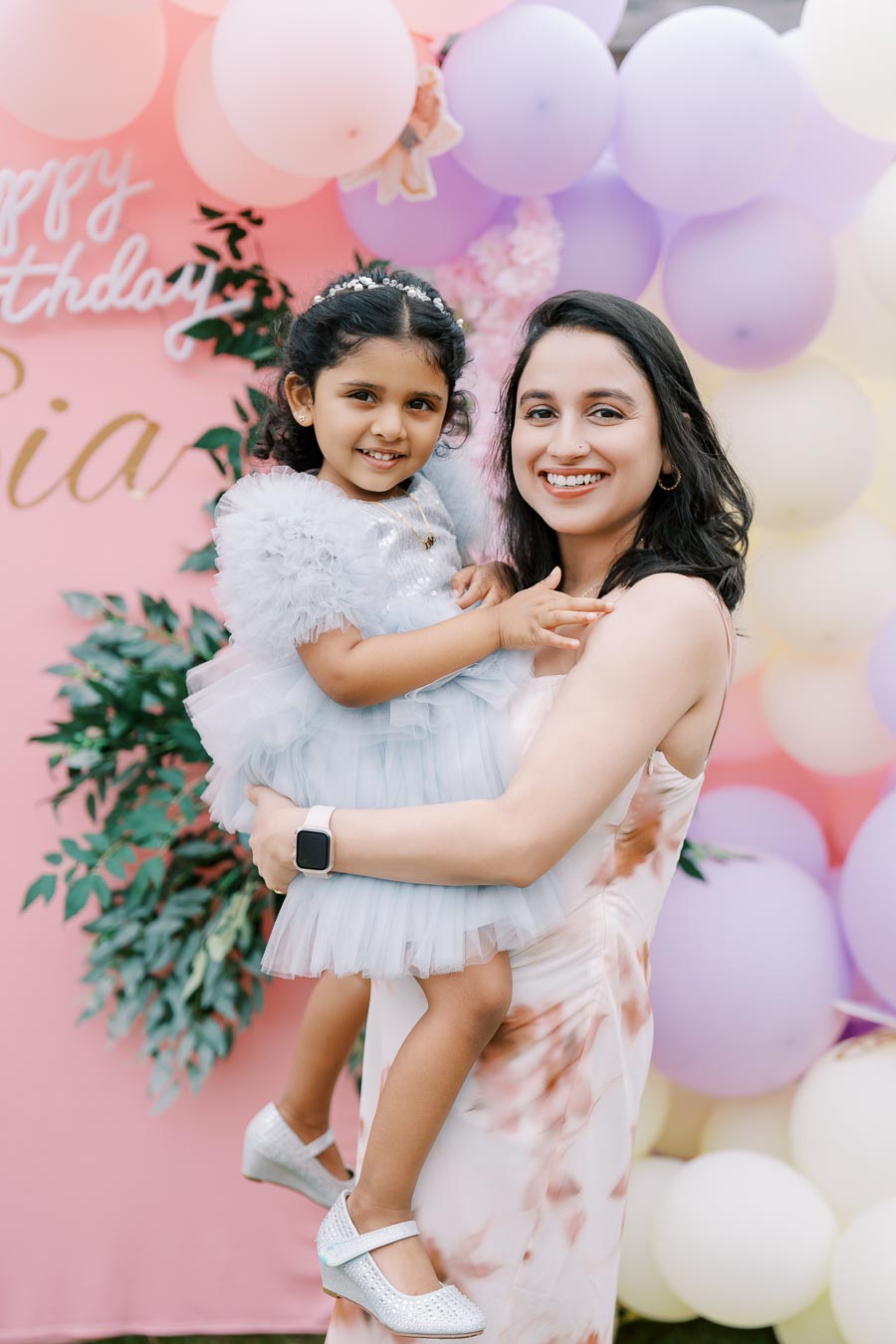 A smiling woman holding a young girl at a birthday party, surrounded by colorful balloons and a decorative background.