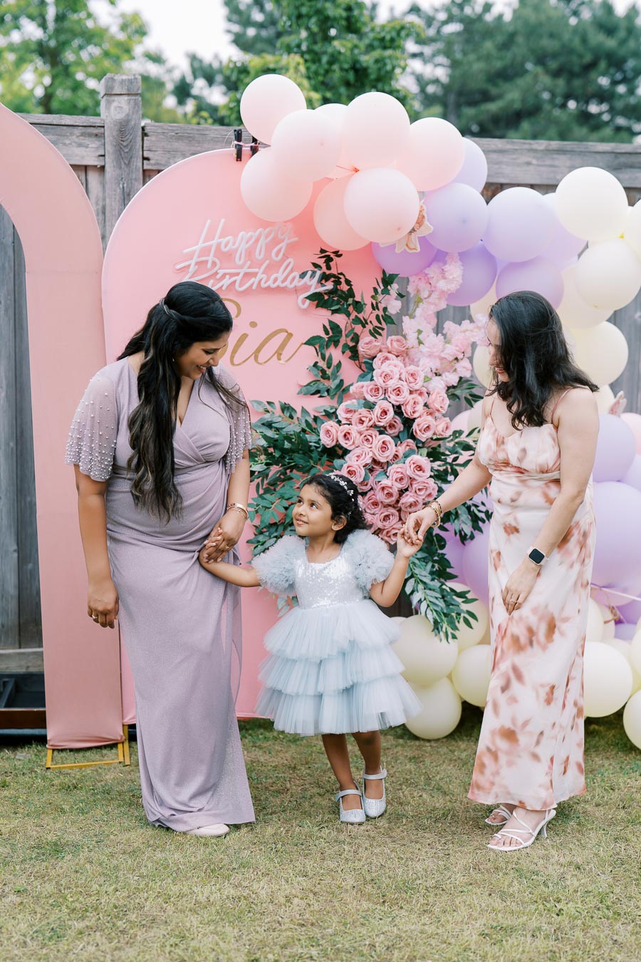 A joyful outdoor birthday celebration featuring two women and a young girl in elegant dresses, standing in front of a decorative backdrop with pink, purple, and white balloons and a Happy Birthday sign, surrounded by pink roses.