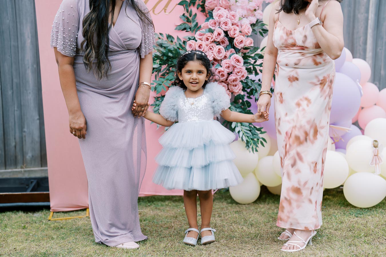 A young girl in a fluffy blue dress holds hands with two women in elegant dresses, standing in front of a festive backdrop with pink roses and balloons.