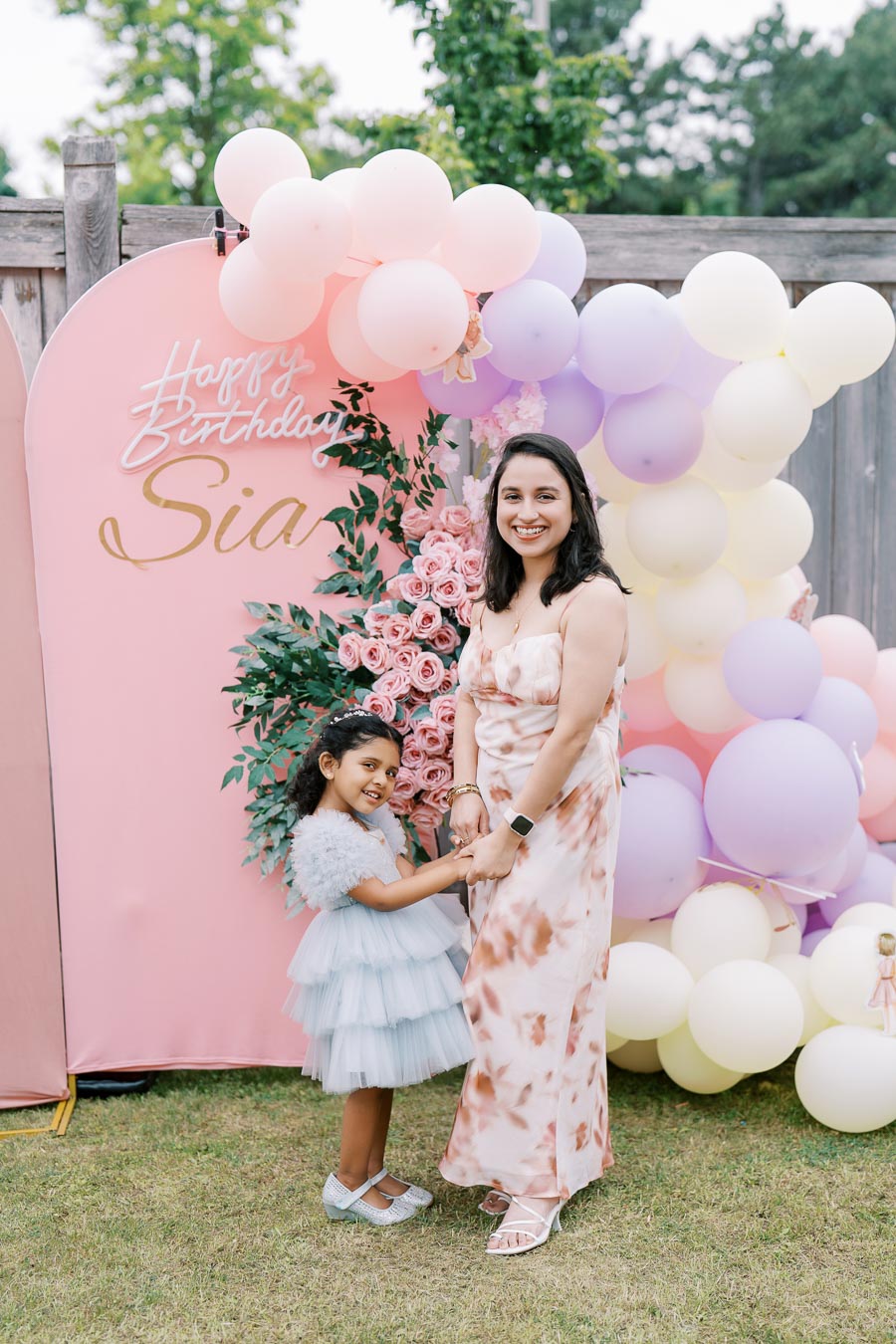 A woman and a child pose in front of a colorful birthday backdrop featuring pink, purple, and white balloons with floral decorations and a Happy Birthday sign.