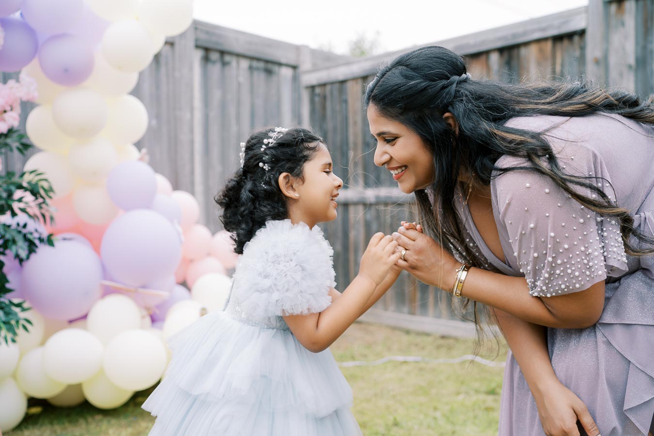 A mother and daughter smiling at each other at a celebratory outdoor event, surrounded by pastel-colored balloons. The little girl wears a fluffy white dress, and the mother is in a beaded, light purple outfit.