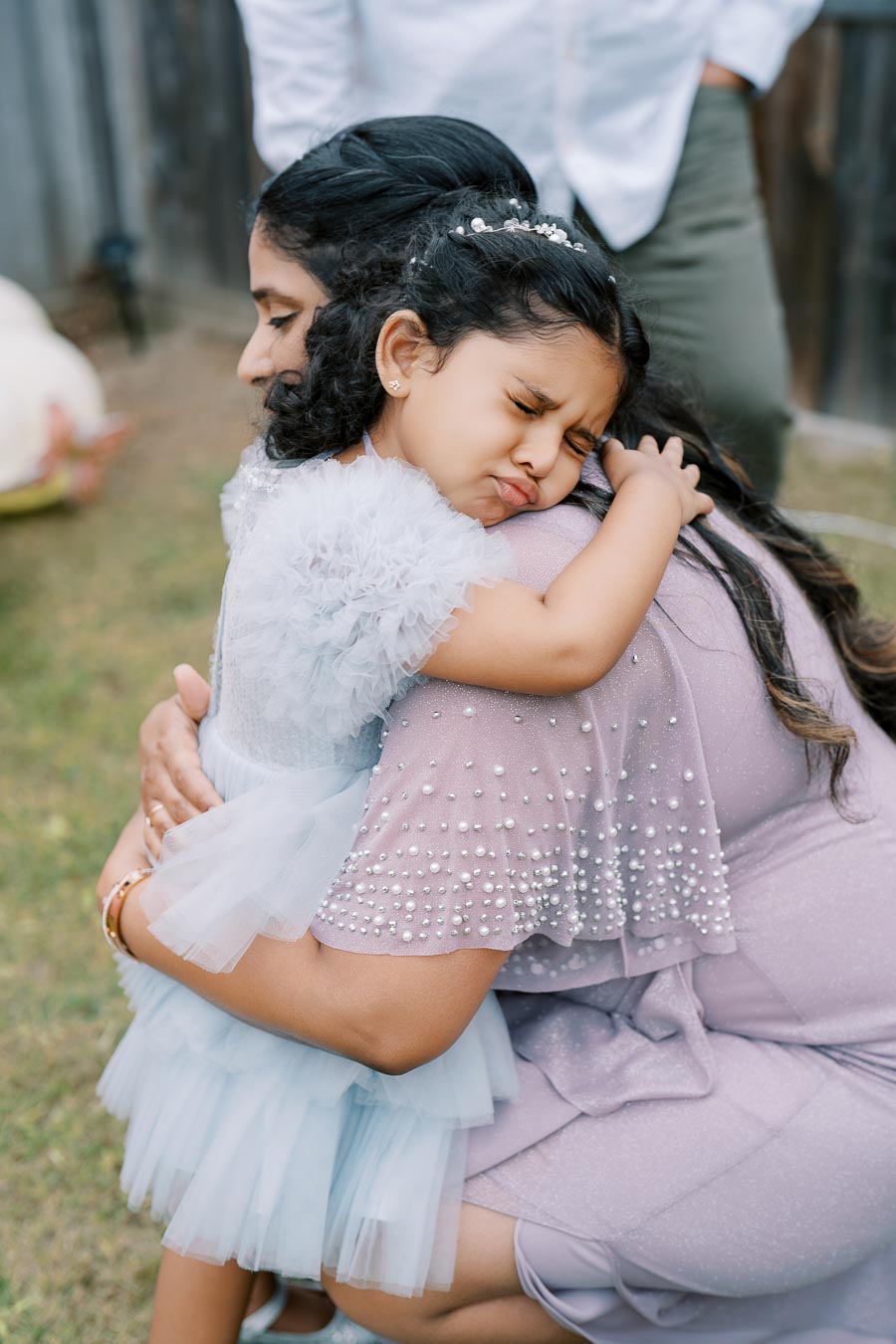 A young girl in a light blue dress hugs a woman in a purple dress adorned with pearls, expressing a tender moment outdoors.