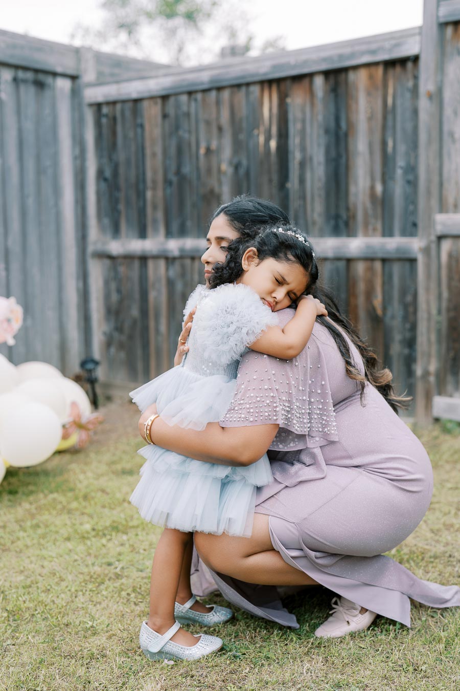 A mother and daughter in elegant dresses share a loving hug in a backyard with a wooden fence and balloons in the background, conveying warmth and familial affection.