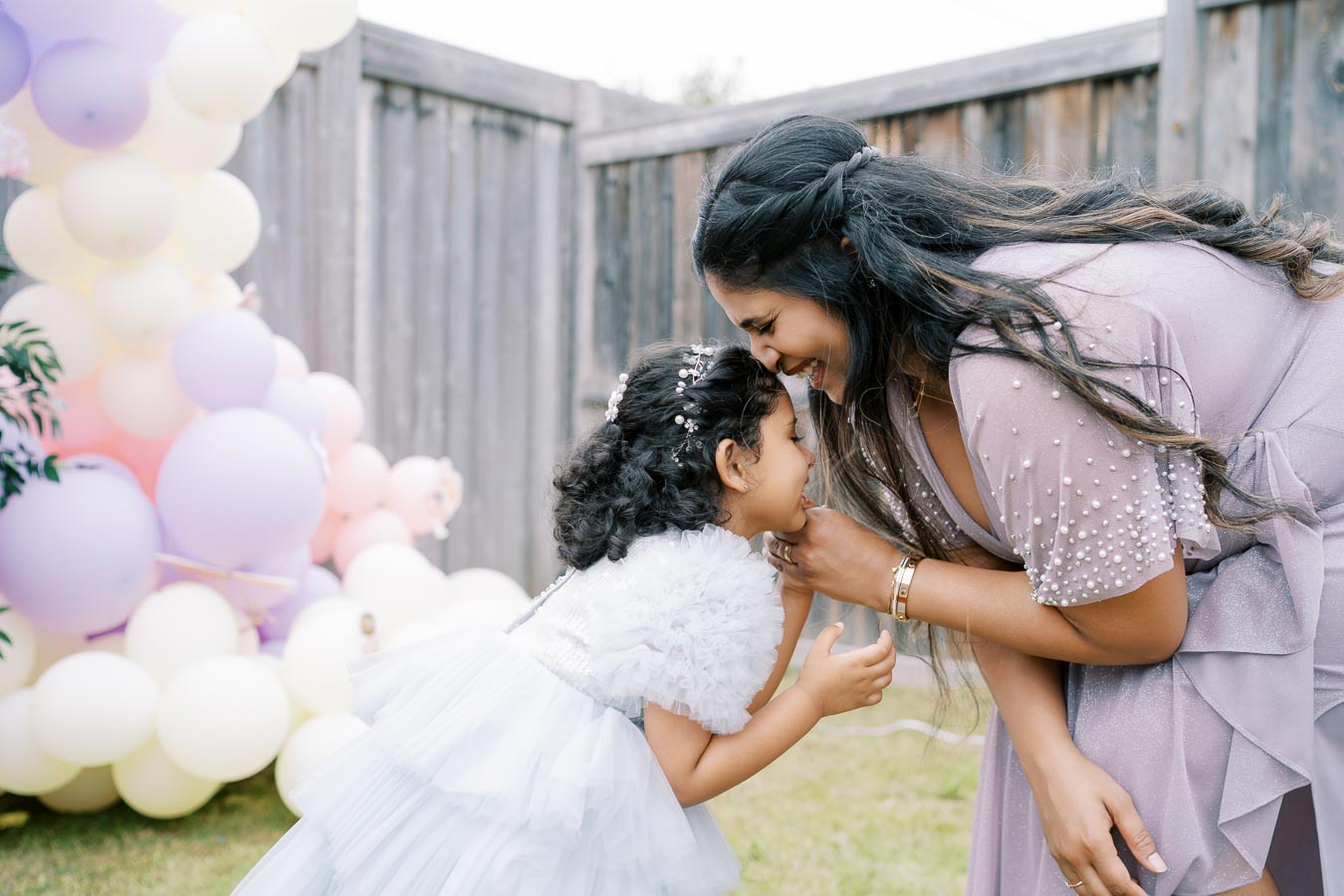 A mother and daughter sharing a joyful moment at a backyard birthday party, surrounded by pastel balloons and wearing elegant dresses.