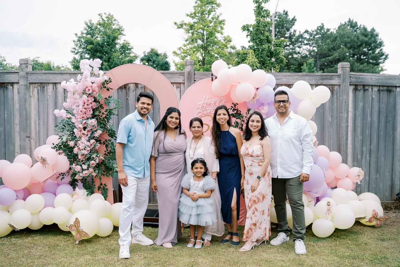 A group of people smiling at a birthday celebration, standing in front of a decorative backdrop with pink and white balloons, pink roses, and a butterfly theme.