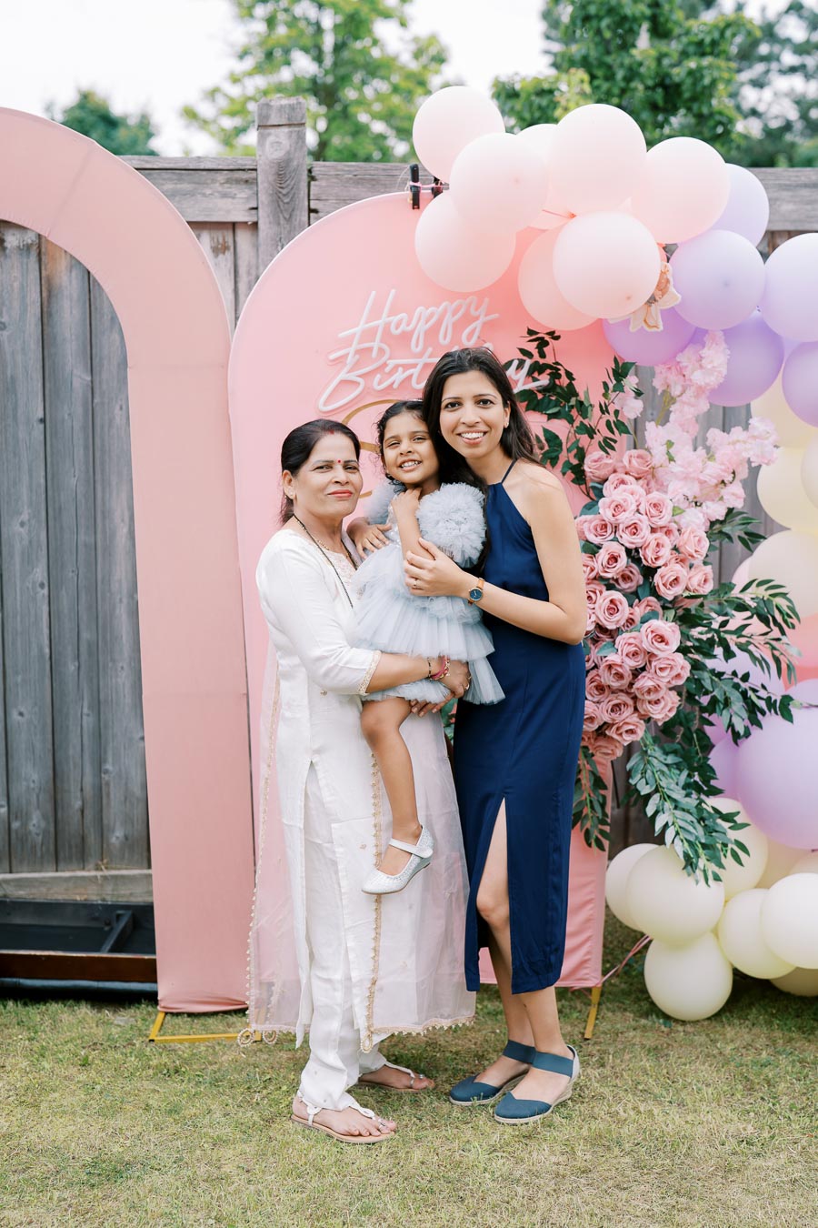 Three women smiling at a birthday celebration with a colorful balloon and floral backdrop.