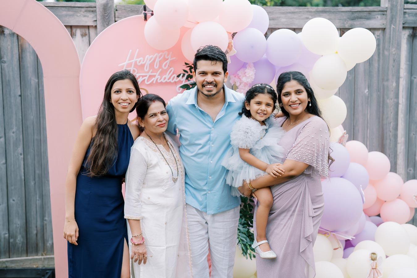 A family celebrating a happy birthday outdoors, with a group of five people smiling in front of a colorful balloon arrangement and a Happy Birthday sign.