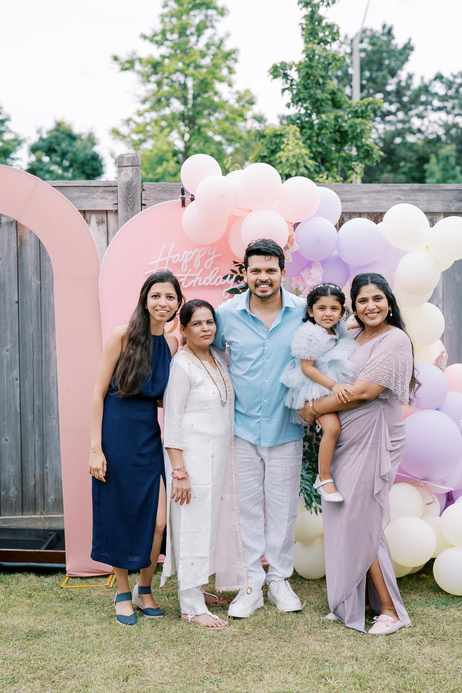 A family celebrating a birthday outdoors, posing in front of a colorful balloon backdrop with a Happy Birthday sign, wearing festive attire.