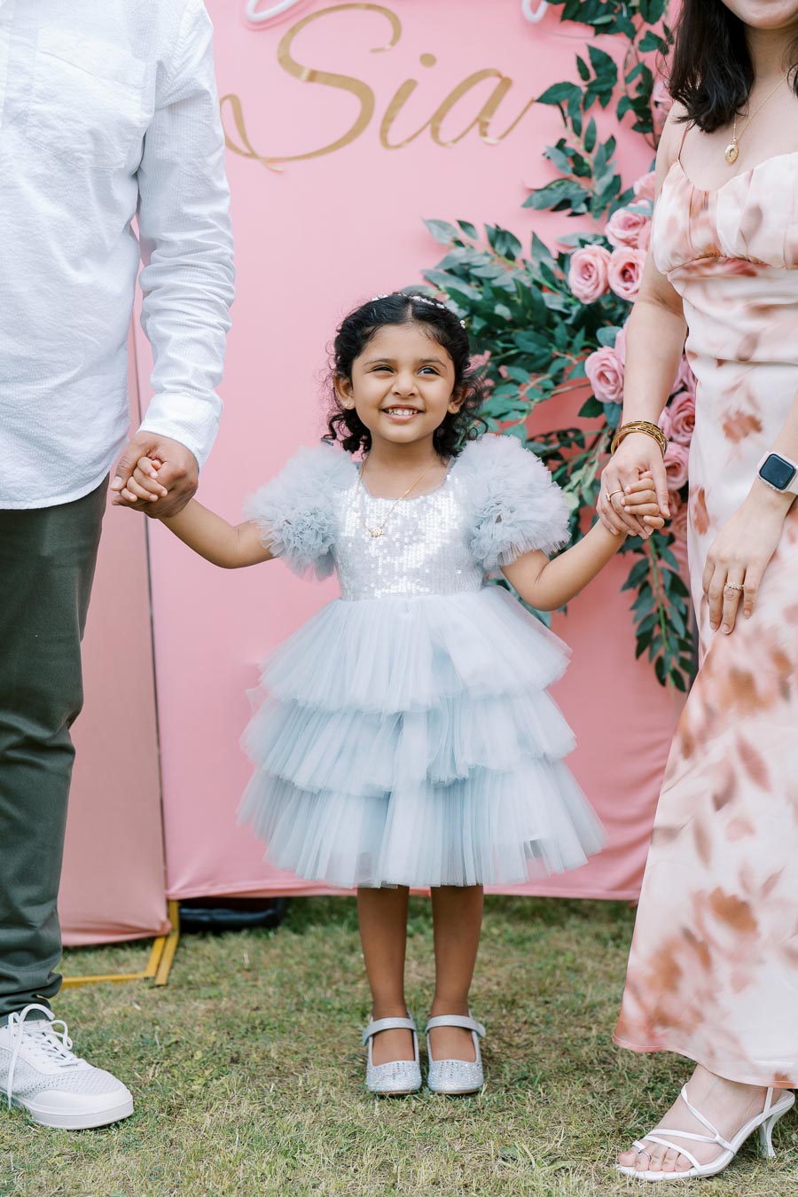 Young girl in a sparkling, blue tiered dress holding hands with adults, celebrating at a pink floral-themed event.